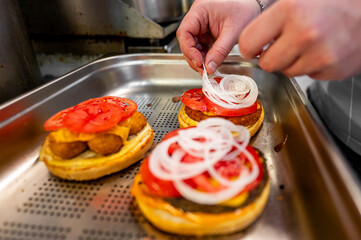 Wall Mural - Close-up of hands placing onion rings on a sandwich with tomatoes and cheese, suggesting food preparation in a kitchen. Ideal for culinary, cooking, and food-related themes.