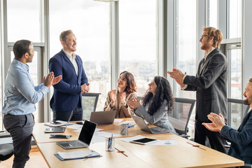 Aufkleber - A business team is gathered around a conference table in a modern office with large windows. They are all clapping and smiling as they applaud a colleague who is standing in front of them.