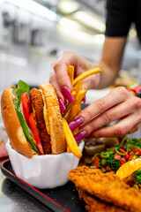 Wall Mural - A close-up of a hand with purple nail polish holding a burger with fries on a kitchen counter. The burger is garnished with fresh lettuce and tomato, showcasing a delicious plant-based meal.