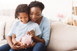 © Prostock-studio - Money Saving Concept. Excited black mom and daughter putting coins into piggy bank, free space