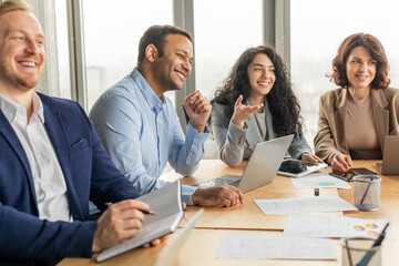 Aufkleber - A group of business professionals, including two men and two women, participate in a meeting around a long wooden conference table. There are multiple laptops, papers, and tablets present