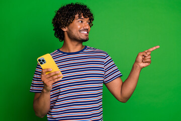 Poster - Portrait of young curly haired guy in striped t shirt using smartphone direct finger mockup interested isolated on green color background