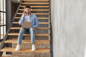 Aufkleber - A man sits on a wooden staircase with headphones on, working on a laptop while smiling. He is wearing blue jeans and a light blue button-down shirt, copy space