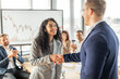 © Prostock-studio - A smiling businesswoman in a grey blazer shakes hands with a colleague in a blue suit after a presentation in an office setting. Other colleagues in the background applaud the businesswoman