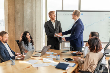 Poster - A group of business professionals are gathered around a large conference table. Two men in suits are shaking hands while standing, signaling a successful agreement or partnership