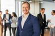© Prostock-studio - A man in a blue suit and white shirt stands in an office setting, smiling confidently. He is surrounded by colleagues, who are talking and working in the background. The office is bright and modern