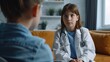 © vefimov - A young girl in a white lab coat sits on a couch with another girl. Scene is serious and focused on the doctor-patient interaction