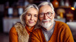 © bluecreamy - retired elderly happy senior couple with a smile, sitting together indoors, in orange sweaters. The man has a white beard, glasses, and the woman has long white hair, in a warm and inviting atmosphere