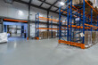 © alhim - Spacious Warehouse Interior With Stacked Pallets and Bright Lighting. The expansive interior of a warehouse showcases organized stacks of pallets along orange and blue shelving units. A loading area