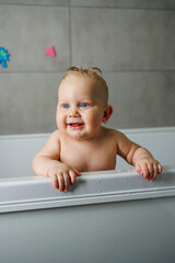 Wall Mural - A blue-eyed baby with a bare torso is standing in the bathroom and smiling. Emotions of joy in a baby. Baby's personal hygiene.