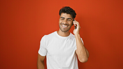 Wall Mural - A cheerful young hispanic man with a beard talking on the phone against a red background