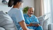 © The Little Hut - Nurse senior and discussion with tablet on sofa for healthcare communication or consultation at home Woman or medical caregiver helping and talking to elderly male person on technology : Generative AI