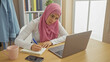 © Krakenimages.com - Focused young woman wearing a pink hijab working on laptop at home, with notebook and smartphone on desk.