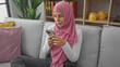 © Krakenimages.com - A woman in hijab engrossed in her smartphone while sitting on a sofa in a cozy living room setting