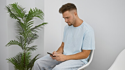 A young hispanic man with a beard wears casual attire while sitting and using a smartphone in a well-lit indoor room with a plant.