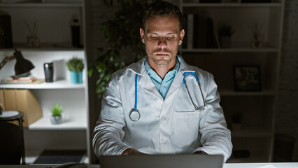 Wall Mural - Focused hispanic man with a beard wearing a lab coat and stethoscope working on a laptop in a modern clinic office.