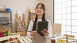 © Krakenimages.com - A young asian woman holds a tablet pensively in a sunny, wood-filled carpentry studio, surrounded by tools and workbenches.