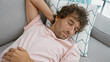 © Krakenimages.com - A young hispanic man in a pink shirt sleeps peacefully on a grey couch, with patterned cushions, in a well-lit, cozy living room.