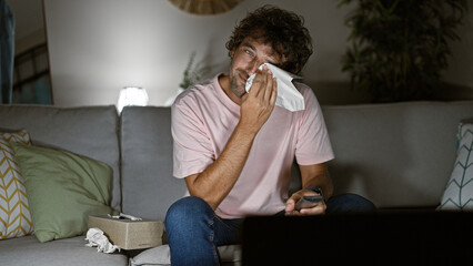 Sticker - Emotional hispanic man crying while sitting on a couch indoors, wiping tears with a tissue.