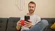 © Krakenimages.com - Handsome middle-aged man with beard lounging on sofa, using smartphone and holding red mug in cozy room.