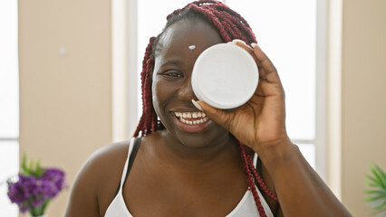 Poster - Happy african woman with braids holding skincare cream in a bright indoor home setting.