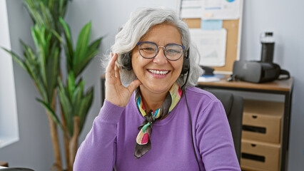 Sticker - A cheerful middle-aged woman with grey hair wearing glasses and a headset smiles in a modern office.