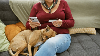 Wall Mural - A middle-aged woman sits on a sofa indoors, using her smartphone with a credit card while a dog rests beside her.