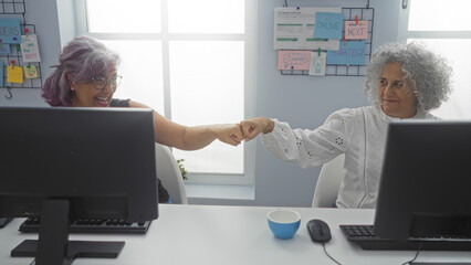 Sticker - Women coworkers in an office celebrating a success with a fist bump, highlighting their teamwork and partnership against a professional and positive workplace backdrop.