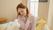 © Krakenimages.com - A young woman with red hair feels neck pain in a cozy bedroom setting, looking down thoughtfully.