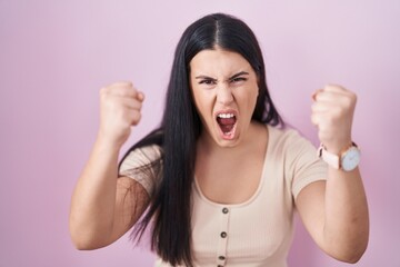 Canvas Print - Young hispanic woman standing over pink background angry and mad raising fists frustrated and furious while shouting with anger. rage and aggressive concept.