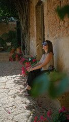 Sticker - Young hispanic woman enjoying a serene moment outdoors in mallorca, spain, seated beside a rustic stone wall adorned with pink flowers and leafy vines.