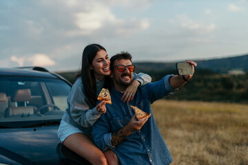 Canvas Print - Loving couple taking a selfie outdoors, leaning on the hood of their car in the evening, eating fast food