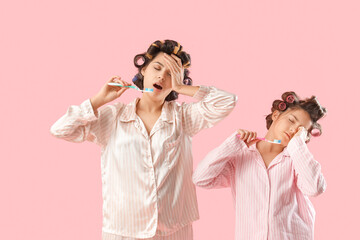 Poster - Sleepy little girl and her mother with hair curlers in pajamas brushing teeth on pink background