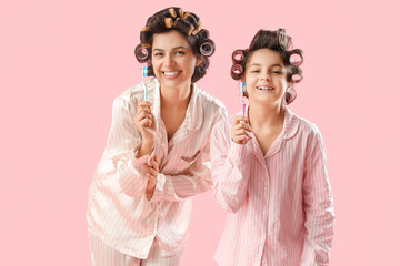 Wall Mural - Little girl and her mother with hair curlers in pajamas brushing teeth on pink background