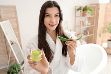 Poster - Beautiful young happy woman with plant leaf and jar of aloe vera gel in bathroom