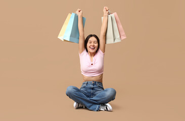 Poster - Fashionable young Asian woman in stylish jeans with shopping bags sitting against brown background