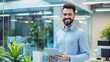© @ArtUmbre - A young man with a beard, wearing a light blue shirt, standing in a modern office setting. He is holding a tablet in his hands and has a cheerful expression on his face