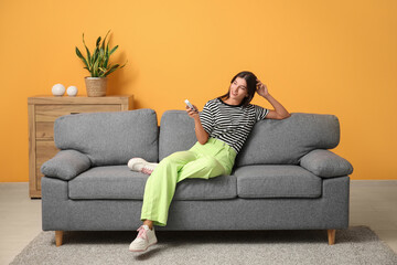 Sticker - Young woman with air conditioner remote control sitting on sofa near orange wall at home