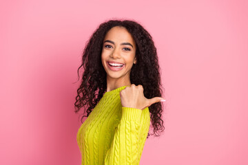 Wall Mural - Portrait of beautiful curly haired brunette girl in green knitted pullover pointing thumb mockup isolated on pink color background