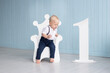 © Marharyta - Little boy on his first birthday sits on a throne-shaped chair, with the number one next to him, dressed in a suit with trousers on pull-ups and a shirt near blue wall at home