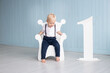 © Marharyta - Little baby boy on his first birthday sits on a throne-shaped chair, with the number one next to him, dressed in a suit with trousers on pull-ups and a shirt near blue wall at home