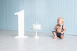 © Marharyta - Little baby boy on his first birthday dressed in trousers on pull-ups and the bow tie sitting on floor next to the cake on the stand, with the number one next to him, smash cake party