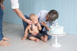 © Marharyta - Mother and her son little baby boy on his first birthday dressed in trousers on pull-ups and the bow tie sitting on floor next to the cake on the stand, with the number one next to him