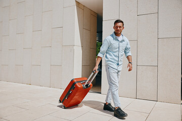 Young man in casual attire confidently walking with a red suitcase outside a modern building