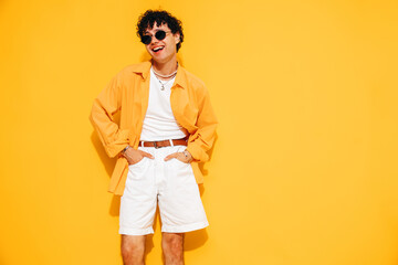 Wall Mural - Young attractive man with short curly hair hairstyle. Smiling handsome male in casual stylish shirt and shorts clothes posing in studio, isolated on yellow. Cheerful and happy model, sunglasses