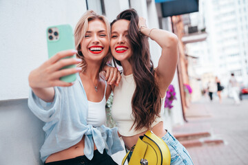 Wall Mural - Two young beautiful smiling female in trendy summer clothes. Sexy carefree women posing in the street. Positive models having fun, hugging in sunny day. With bright makeup and red lips, take selfie