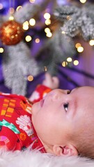 Wall Mural - Healthy little boy in festive red clothes lies under the Christmas tree. Baby looking straight ahead. Blurred backdrop. Vertical video