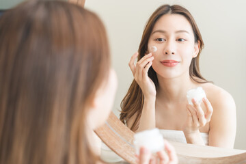 Wall Mural - Facial beauty skin care, smile of pretty asian young woman in bathrobe looking at mirror, hand applying moisturizer lotion on her face, holding jar of skin cream before makeup cosmetic routine at home