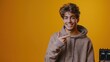 © MakoPoko - Young smiling man in a hoodie pointing with his thumb to a mockup workspace, isolated on a bright yellow-orange background.