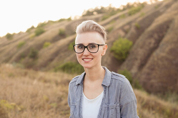 Woman with short haircut hairstyle in glasses wearing blue shirt in nature outdoors on sunset near green hills